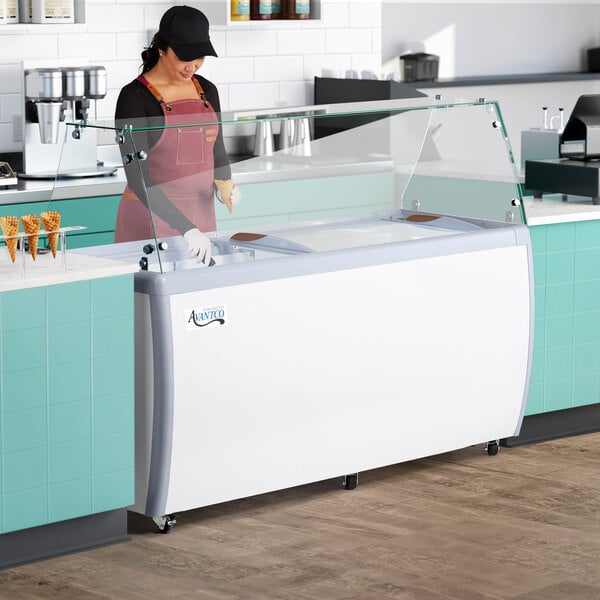 An aproned woman in gloves standing in front of an Avantco gelato dipping cabinet.