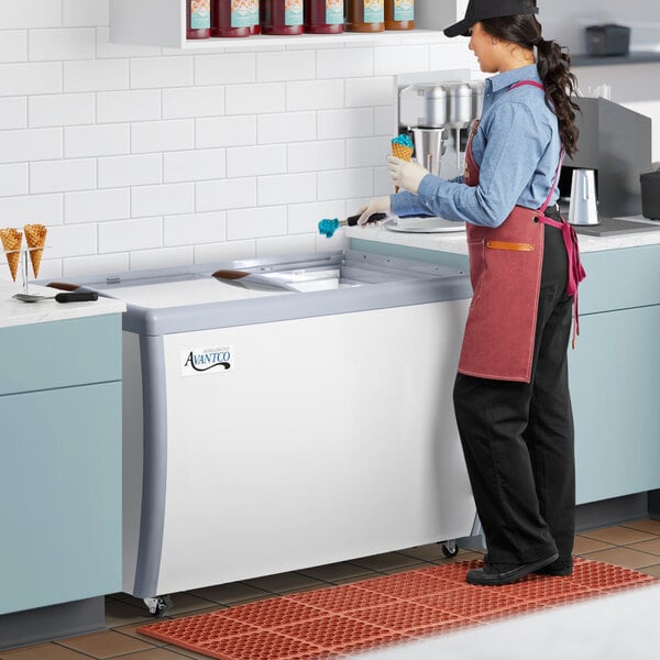 An aproned woman standing in front of a Avantco gelato dipping cabinet on a counter.