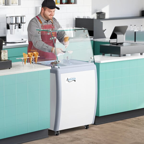 A man wearing a black cap and apron holding an ice cream cone in front of a white and grey Avantco gelato dipping cabinet.