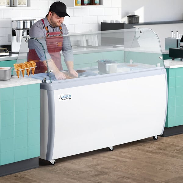 An Avantco gelato dipping cabinet on a counter in an ice cream shop with a curved glass top and a man in an apron serving gelato.