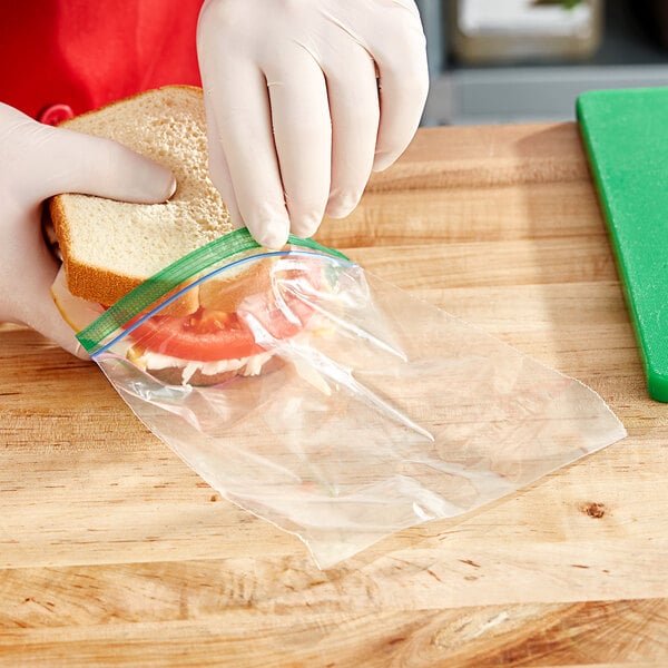 A person in gloves putting a sandwich in a Ziploc sandwich bag on a school kitchen counter.