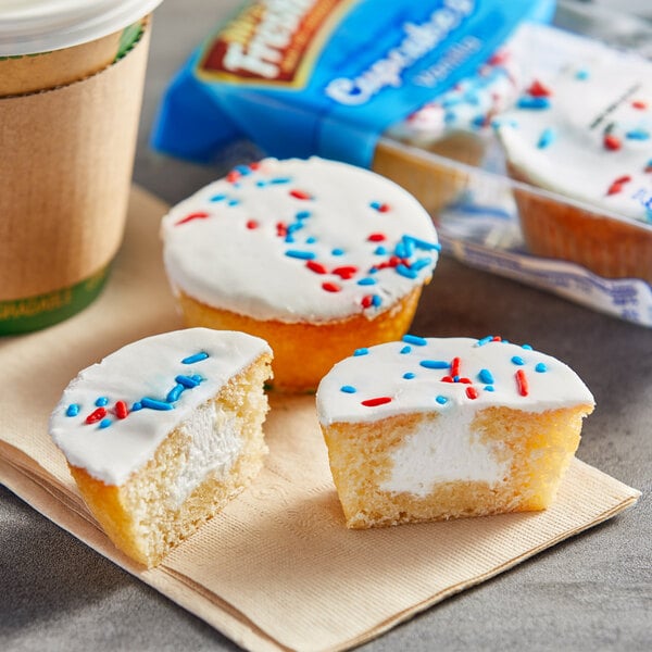 A cardboard container holding three Mrs. Freshley's vanilla cupcakes with white frosting and red and blue sprinkles.