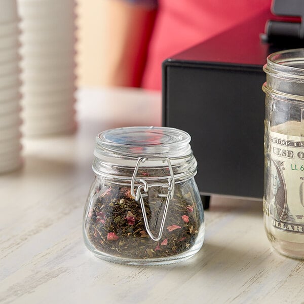 A Choice glass storage jar with a hinge top lid holding a dollar bill and tea.