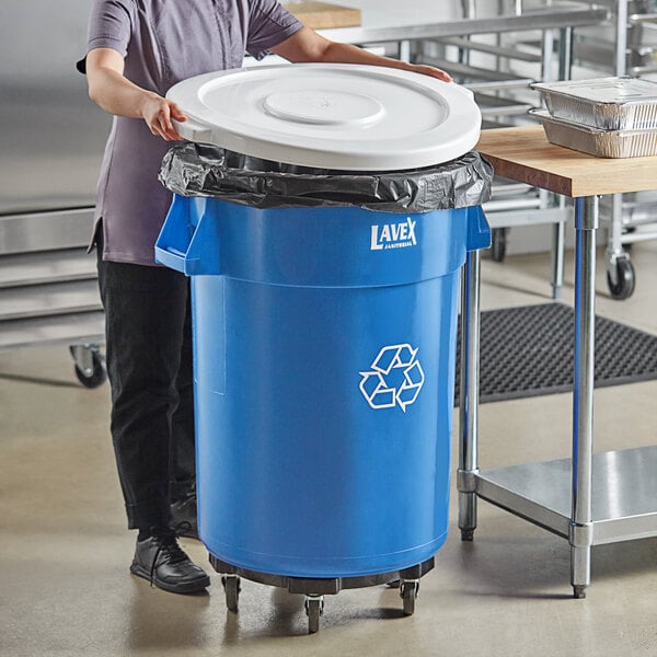 A woman standing next to a Lavex blue round commercial recycling can with a white lid.