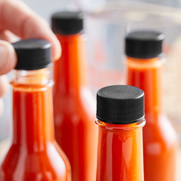 A hand using a 24/414 black plastic lid to close a small bottle of red sauce.