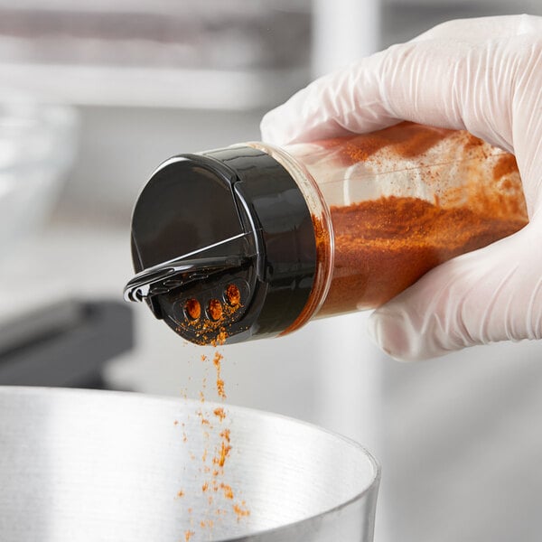 A hand in a glove using a 48/485 black dual flapper spice lid to pour red powder into a container.