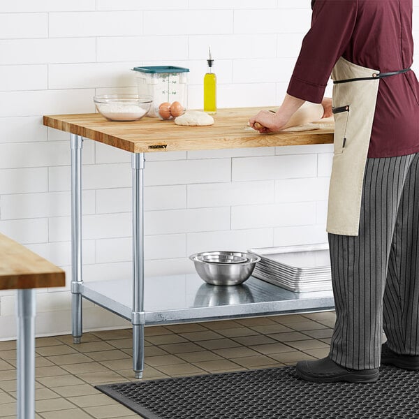A man in a chef's outfit using a Regency wood top work table with a galvanized base and adjustable undershelf to roll dough.