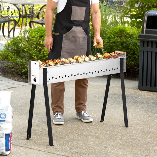 A man wearing an apron cooking on a Backyard Pro stainless steel kebab grill on a table outdoors.