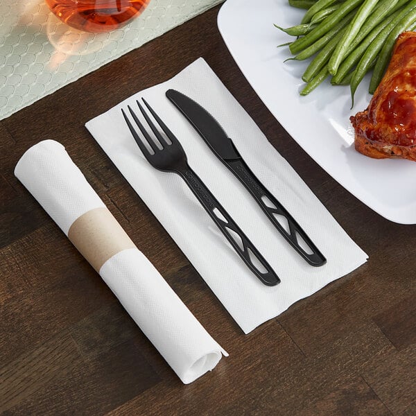 A Hoffmaster FashnPoint white napkin with a pre-rolled black compostable plastic fork and knife set on it.