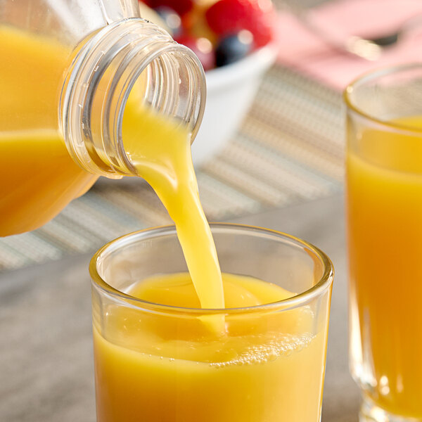 A bottle of orange juice is being poured into a glass, with another filled glass and a bowl of fruit visible in the background.