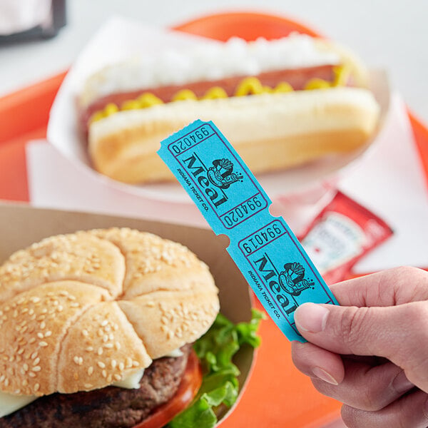 A hand holding a blue "Meal" raffle ticket next to a burger and hot dog.