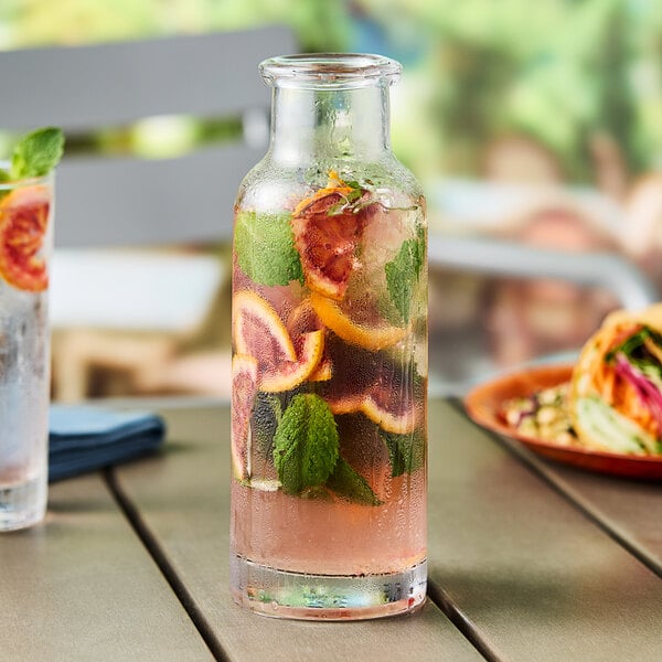 A clear glass Acopa water bottle with a glass of water and orange slices on a table.