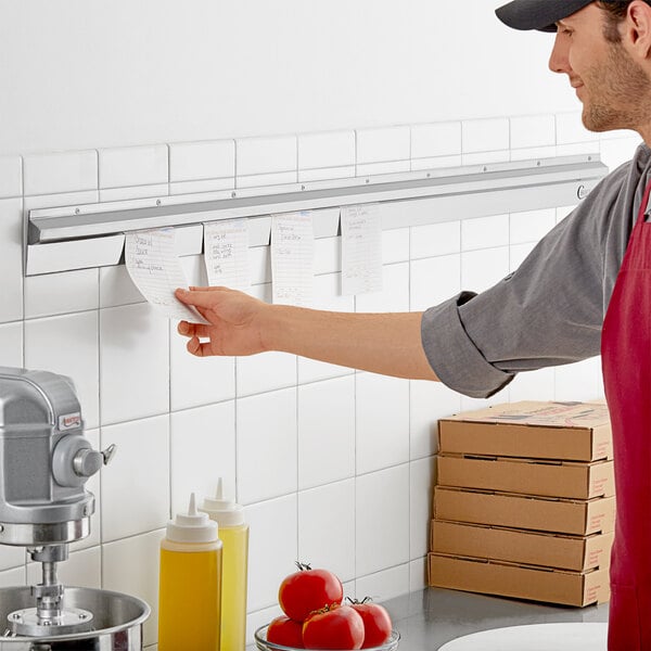 A man in a red apron in a professional kitchen using a Choice stainless steel wall mounted ticket holder.