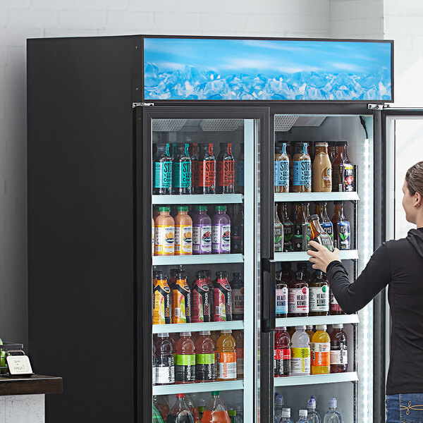 A woman standing next to a refrigerator with a blue sign reading "Beverages" above bottles of drinks.