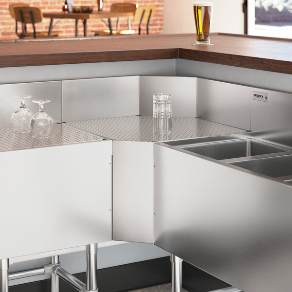 A stainless steel counter with a Regency Spec Line 90-degree angle inside corner underbar filler next to glasses and a glass of beer.