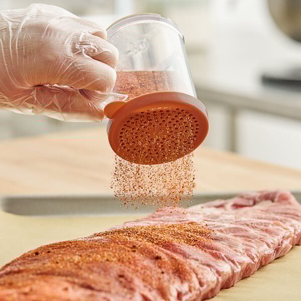 A hand holding a Tablecraft polycarbonate shaker with a red rose lid pouring seasoning onto a piece of meat.