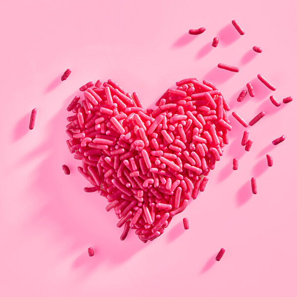 A heart shape made of pink sprinkles on a pink background.