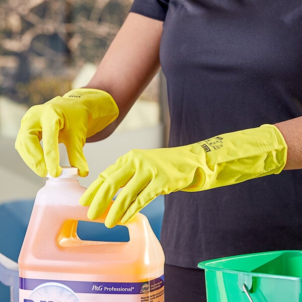 A person wearing Lavex yellow rubber gloves holding a plastic jug of liquid.