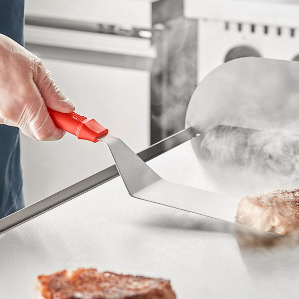 A person holding a Choice 8" x 3" turner with a red handle over meat on a grill.