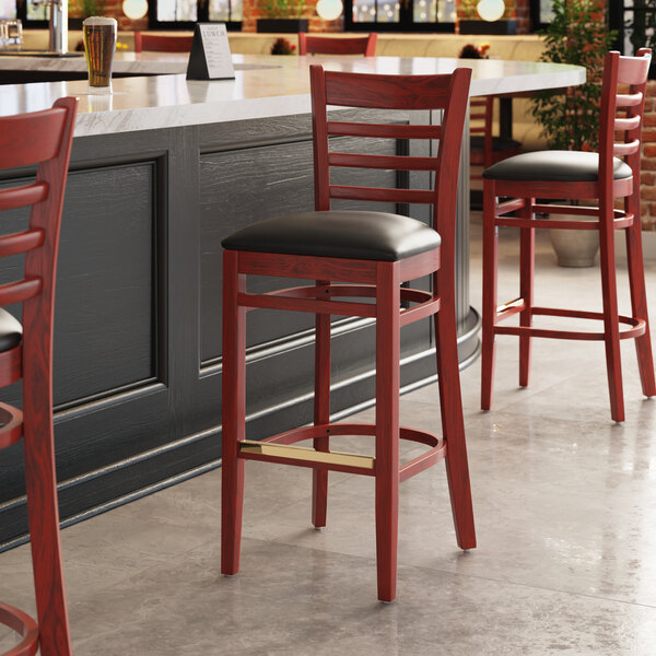 A Lancaster Table & Seating mahogany wood bar stool with black vinyl cushion.