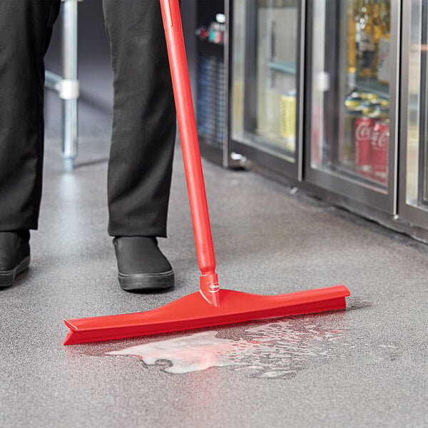 A person using a Vikan red single blade floor squeegee to clean the floor.