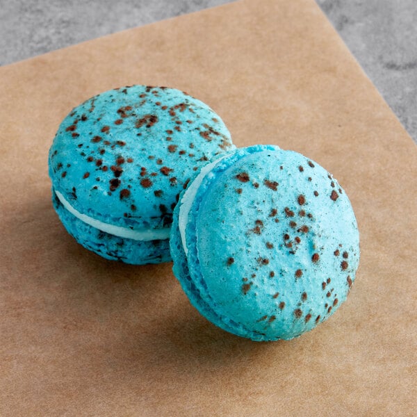 Two blue Macaron Centrale macarons with white chocolate on top and brown speckles.