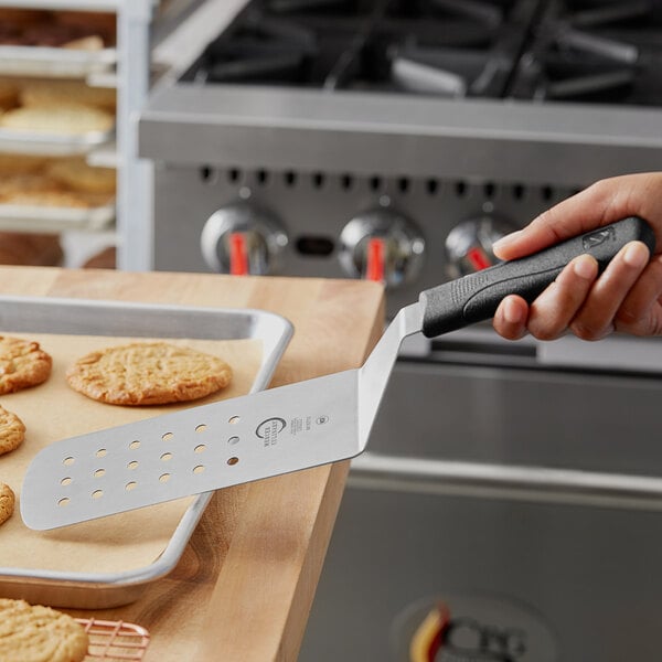 A hand holding a black Mercer Culinary Millennia Perforated Turner over a tray of cookies.