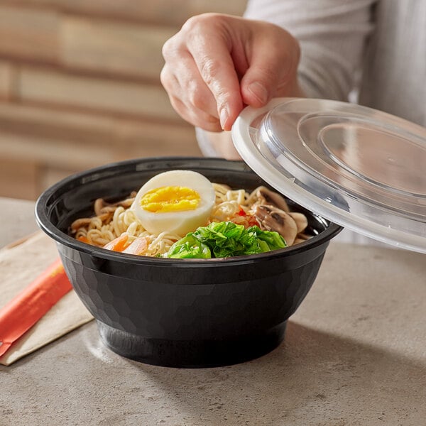 A person holding a Choice black plastic bowl filled with noodles and vegetables with a hard boiled egg.