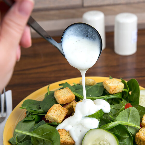 A person using a Fineline black plastic ladle to pour white dressing over a salad.