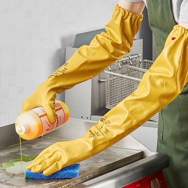 A person wearing Showa yellow rubber gloves cleaning a countertop with a bottle of cleaning liquid.