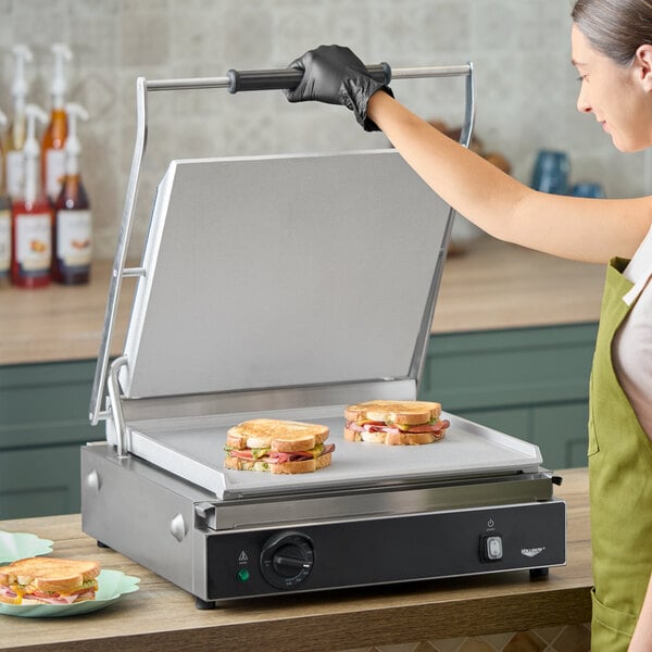 A large stainless steel panini grill with smooth plates, shown open with sandwiches on the cooking surface.