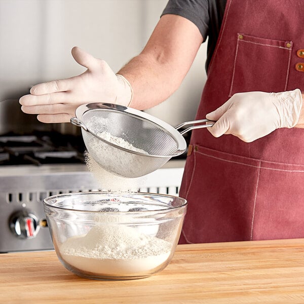 A man using a Matfer Bourgeat stainless steel fine mesh strainer to sift flour into a bowl.