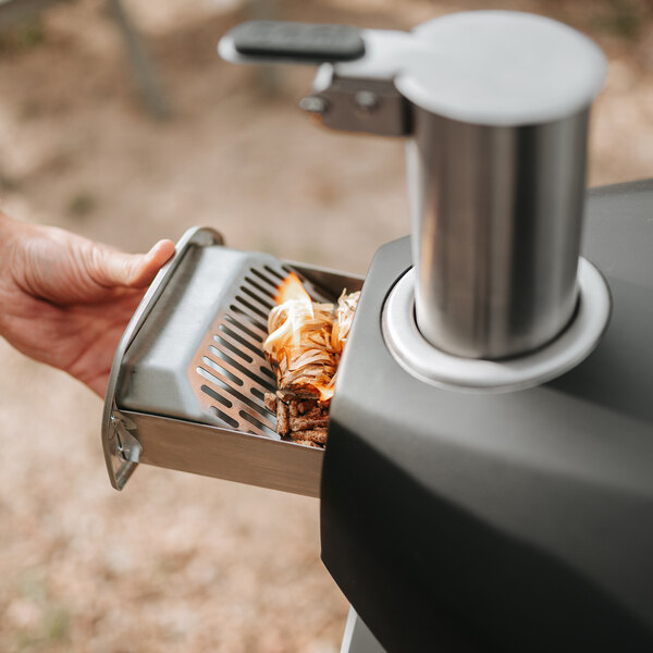 A hand holding a grill with food over a Ooni Fyra outdoor pizza oven.