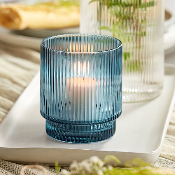 A blue glass Acopa Lore tealight holder with a lit candle on a white tray.