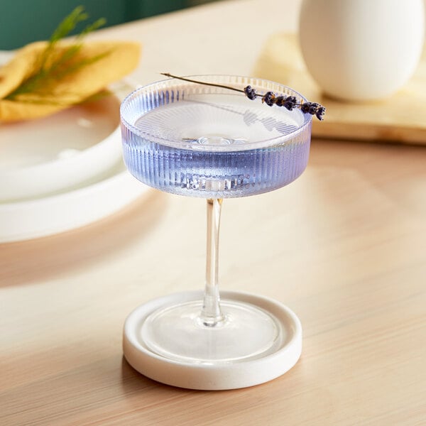 An Acopa coupe glass of blue liquid with a lavender flower in it.