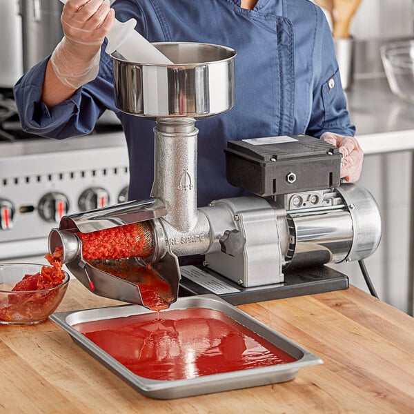 A woman using a Tre Spade Electric Tomato Mill to pour red tomato sauce into a metal container.