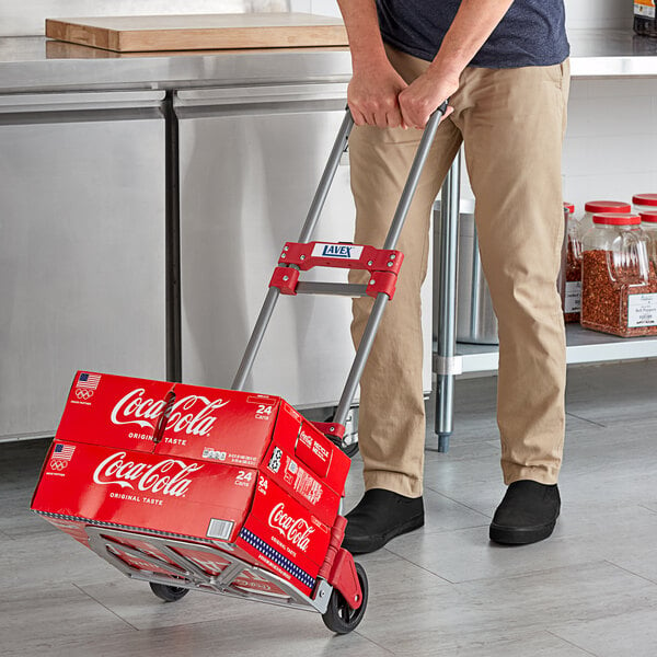 A man pushing a cart with a stack of red boxes of soda on a Lavex Aluminum Folding Hand Truck.