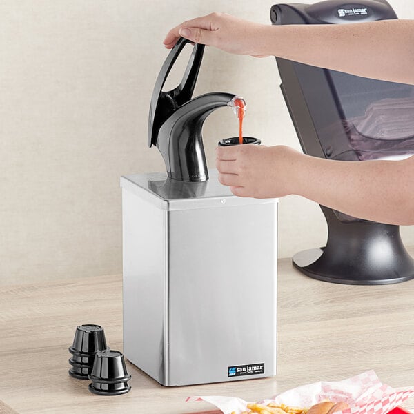A person pouring liquid into a San Jamar countertop condiment dispenser.