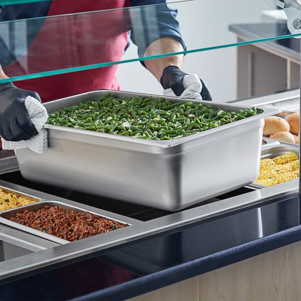 A person in a black glove holding a large stainless steel pan of green beans.