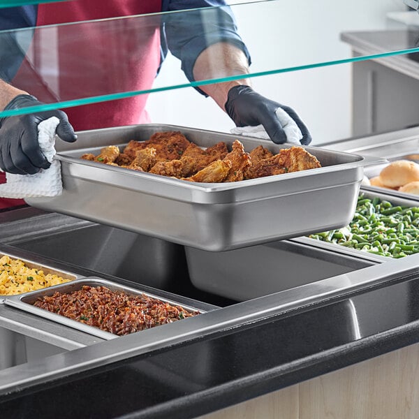 A person in a black apron holding a stainless steel steam table pan of food on a counter.