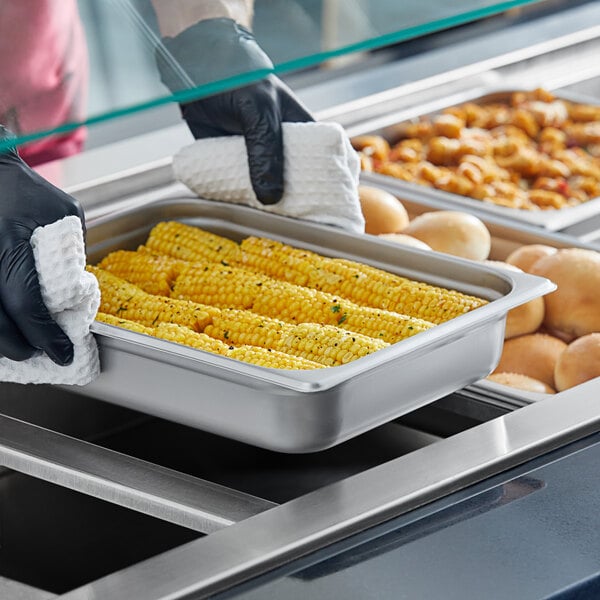 A person wearing black gloves holding a Choice 1/2 Size Stainless Steel Steam Table Pan filled with corn.