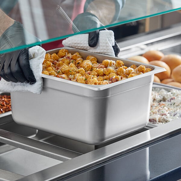 A person in black gloves holding a stainless steel steam table pan over a container of food.