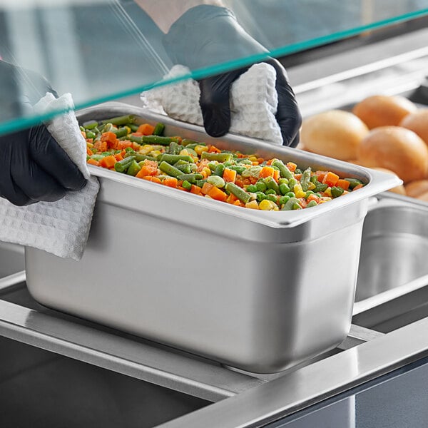 A person holding a stainless steel steam table pan filled with food on a counter.