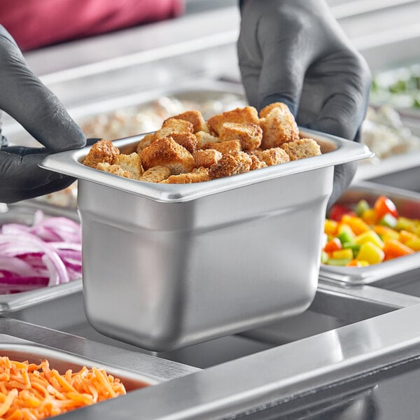 A person in gloves holding a Choice 1/9 size stainless steel steam table pan full of food.