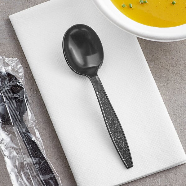 A black plastic spoon wrapped in a plastic bag on a napkin next to a bowl of soup.