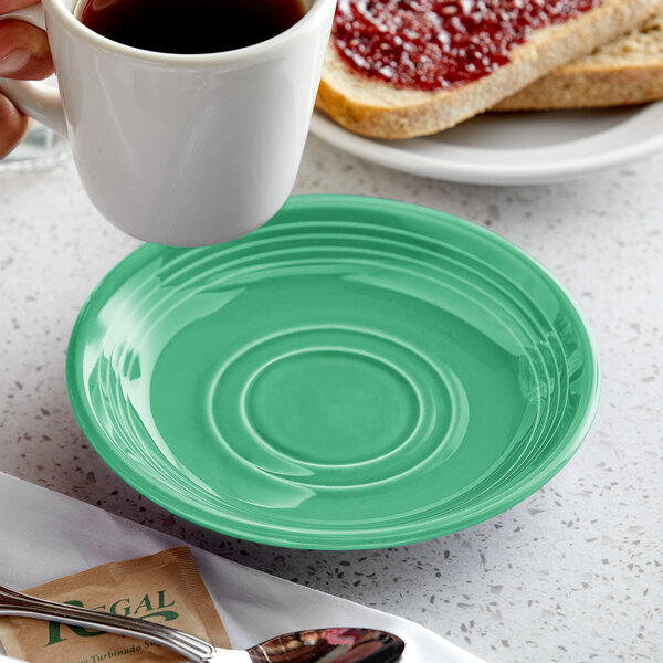 A hand holding a Tuxton Cilantro saucer under a white cup of coffee over a green plate.