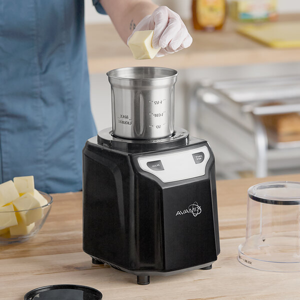 A black AvaMix commercial spice grinder with a stainless steel bowl being loaded with butter in a kitchen setting.