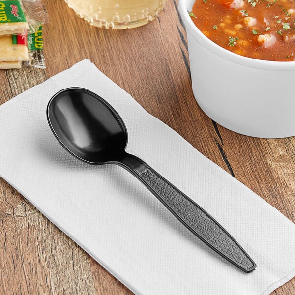 A black Choice plastic soup spoon on a napkin next to a bowl of soup.