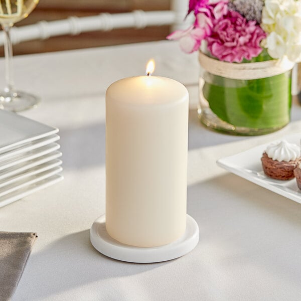 A white Hollowick wax pillar candle on a table with white plates and glasses.