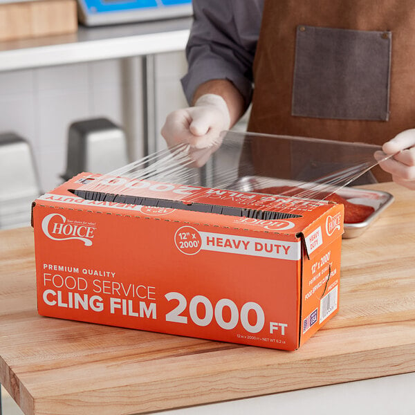 A person using a serrated cutter to cut a box of Choice Heavy-Duty Foodservice Film on a table.